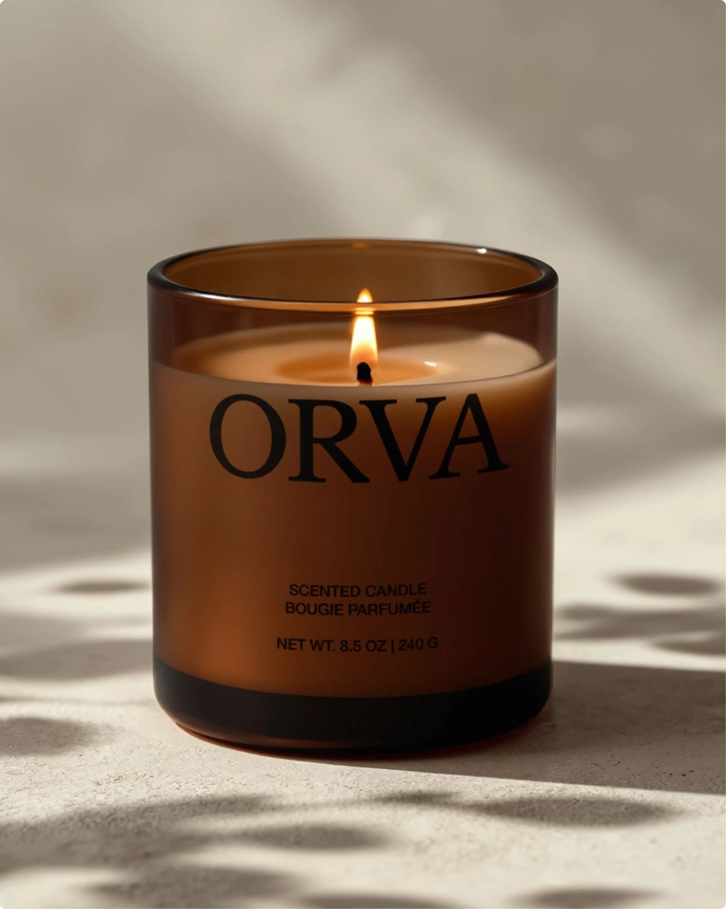 Açık renkli bir yüzeyde yanan kehribar camlı ORVA mumu; kelime markası, SCENTED CANDLE ve BOUGIE PARFUMÉE satırları ile NET WT. 8.5 OZ 240 G okunabiliyor, sert ışık yaprak gölgeleri düşürüyor
